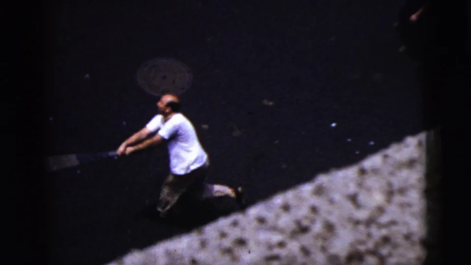 High-angle 1956 New York City street view of an adult man in a white shirt swinging a bat on dark pavement.