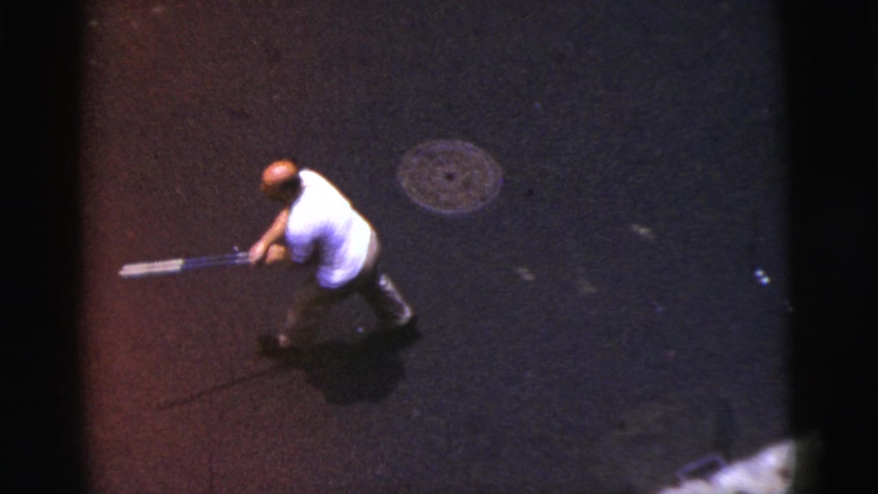 Balding man in a white shirt follows through with a bat on dark pavement.