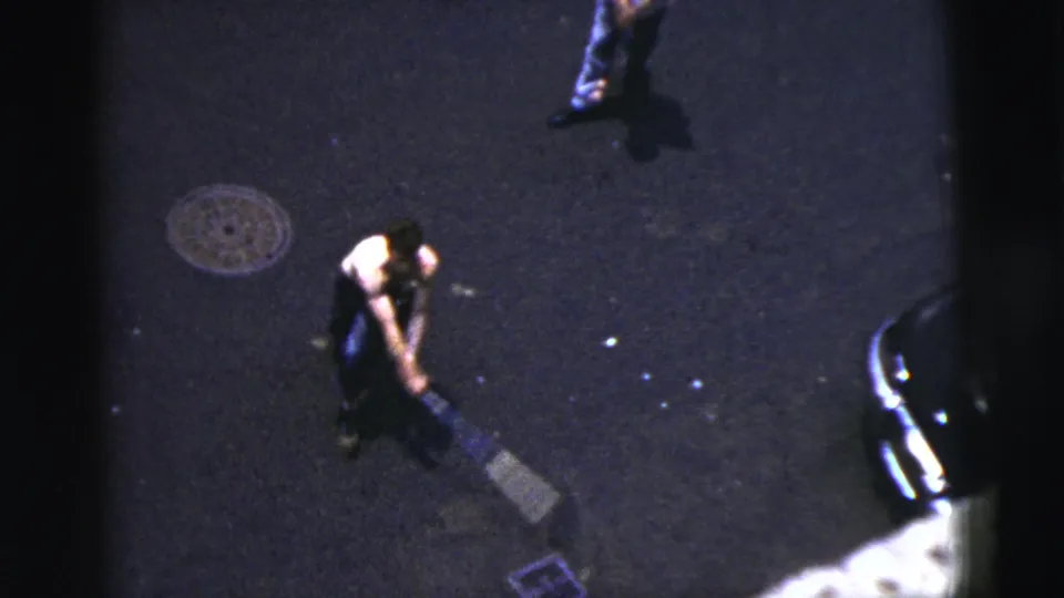 A man swings a bat on a dark New York City street in 1956, with parked-car edges and rough pavement in view.