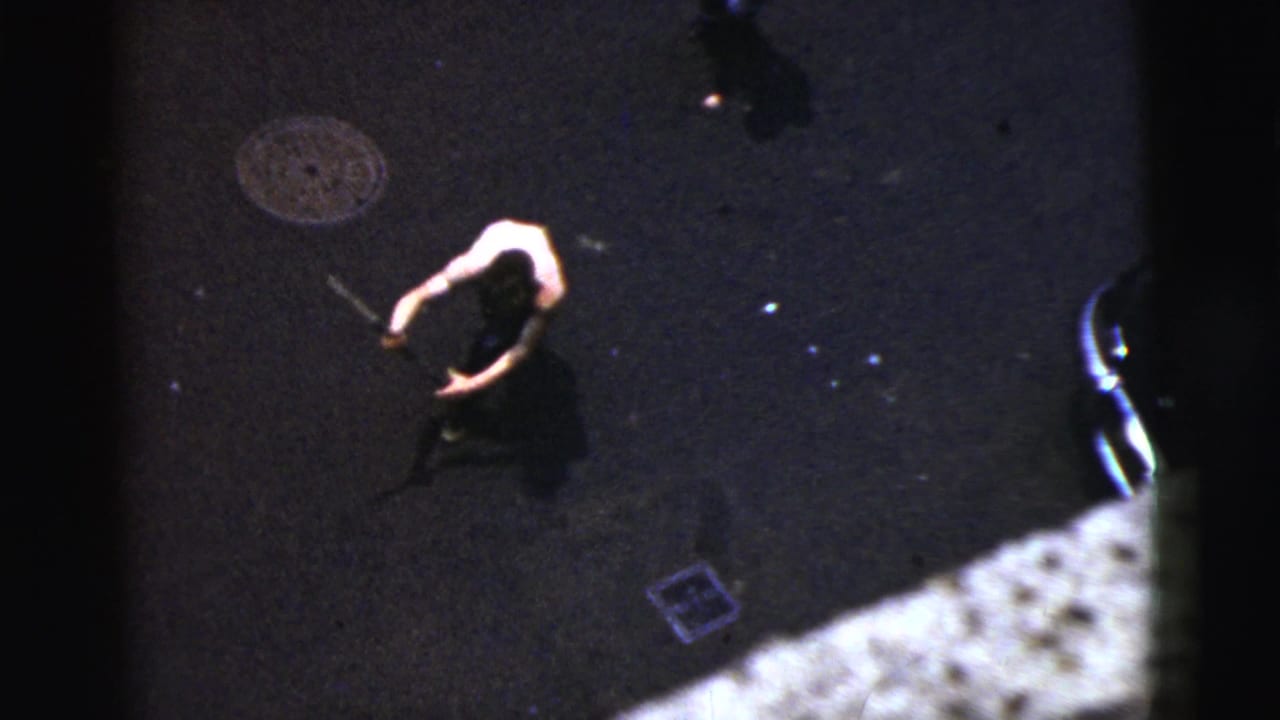 High-angle street view of a person with a bat near a manhole cover and parked car edge.