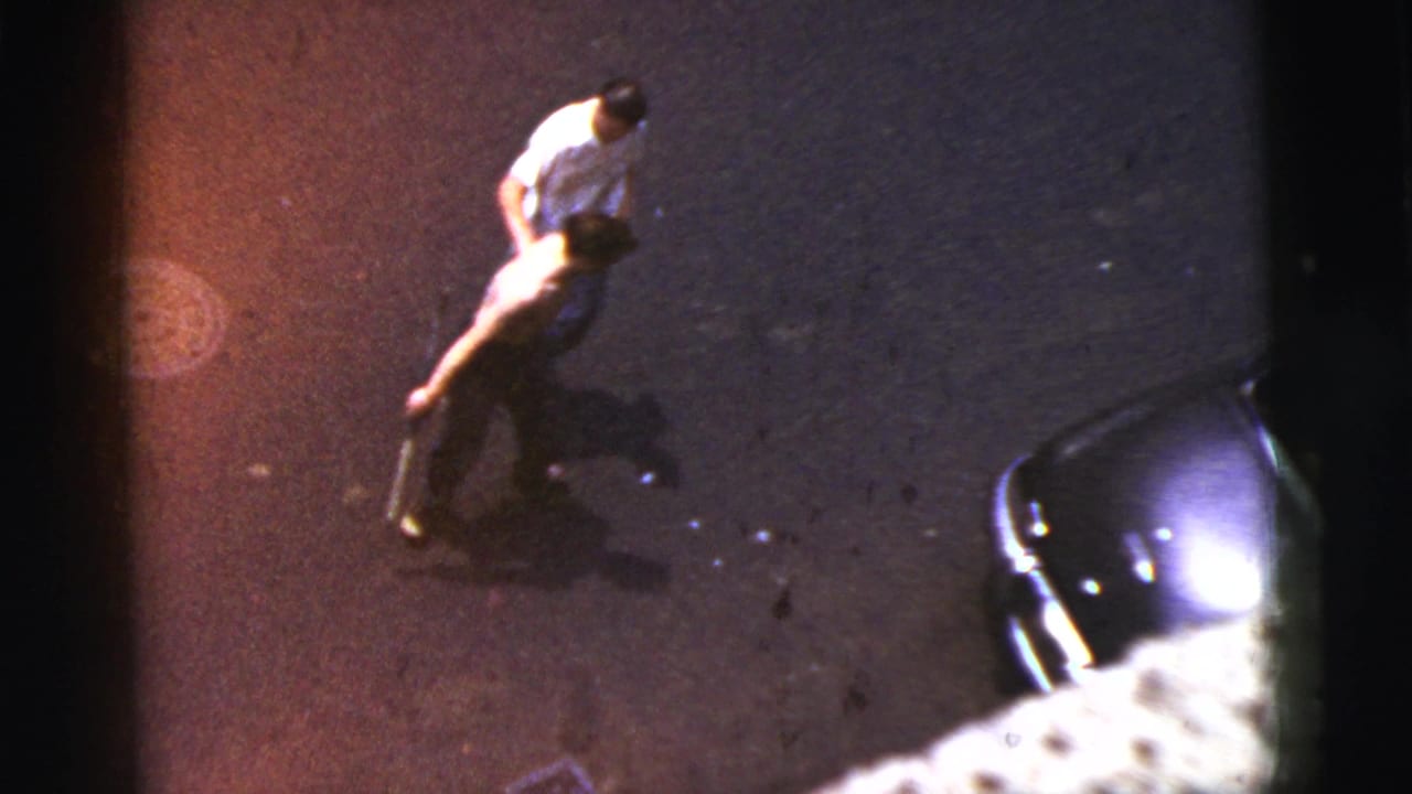 High-angle 1956 New York City home-movie frame of a player swinging a bat on an asphalt street near a parked car.