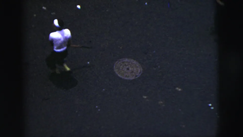 High-angle 8mm frame from 1956 New York City showing a street player from behind with a bat near a manhole cover.