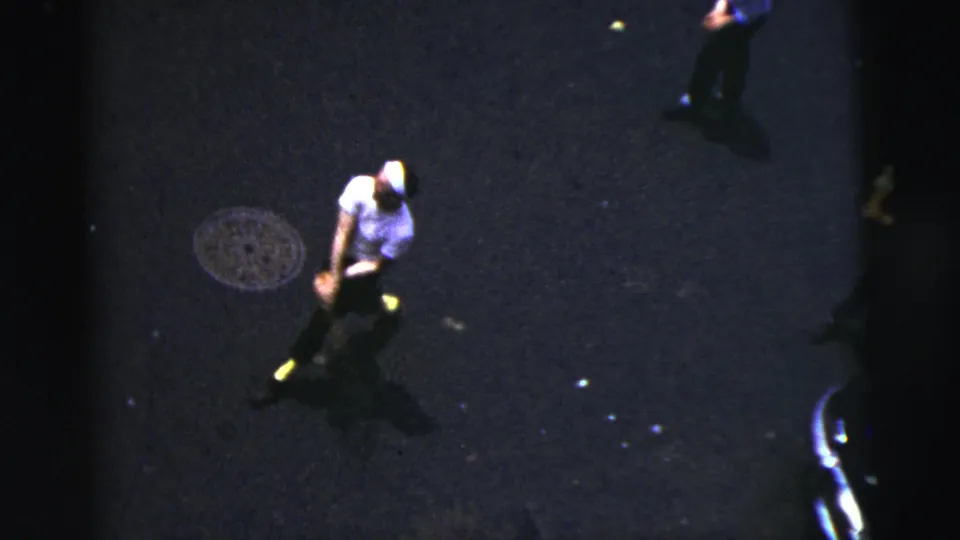 1956 New York City home-movie frame showing a player with a bat on a dark street, with manhole cover and parked car edge visible.