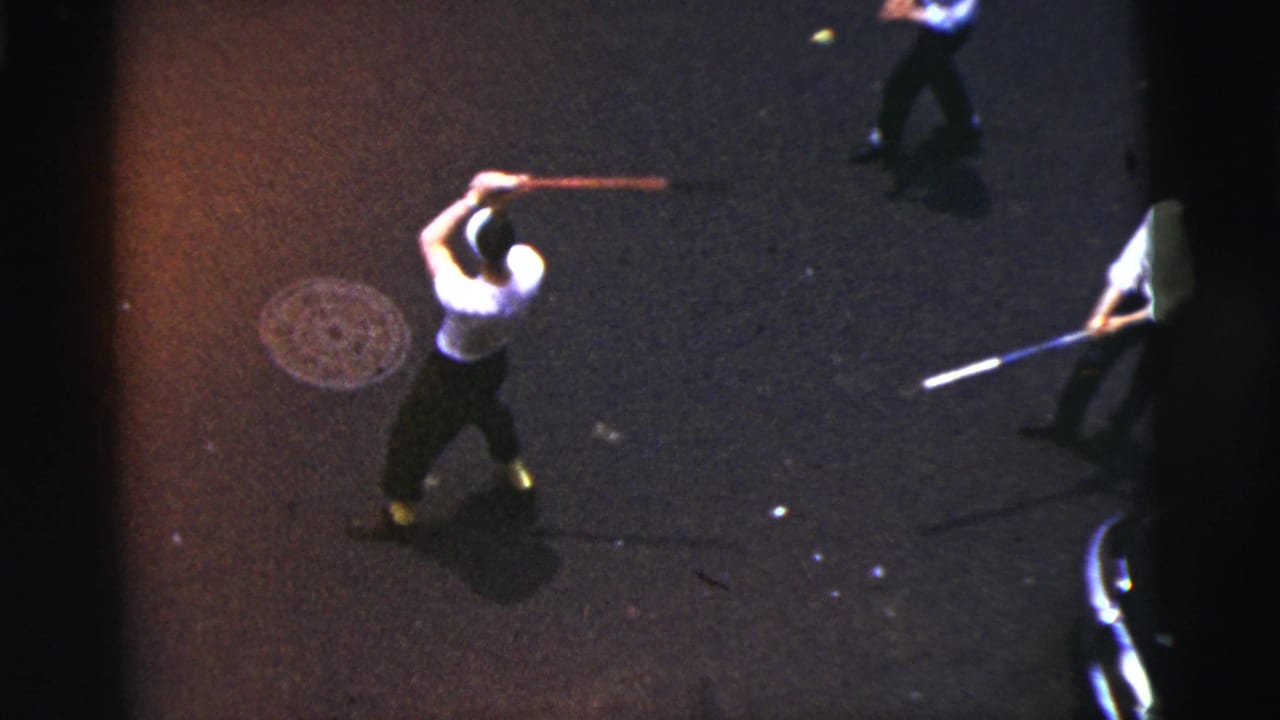 1956 New York City home-movie frame showing a boy in a white shirt swinging a bat on asphalt as others stand nearby.