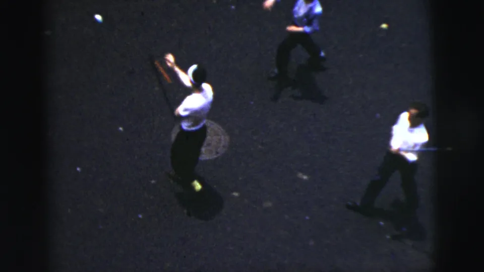 Home-movie frame of boys playing street baseball in New York City, with a raised bat and a manhole cover visible on the asphalt.