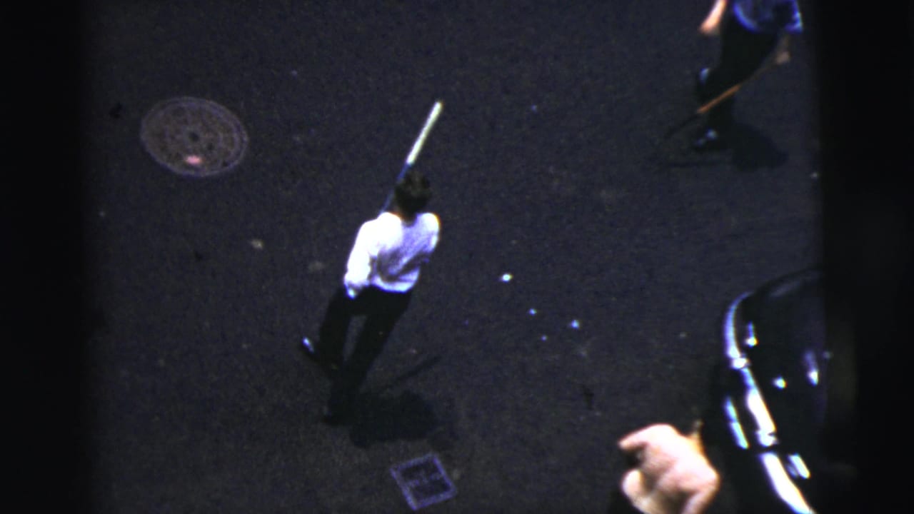 Elevated 1956 New York City 8mm view of a man in a white shirt holding a bat on a dark asphalt street.