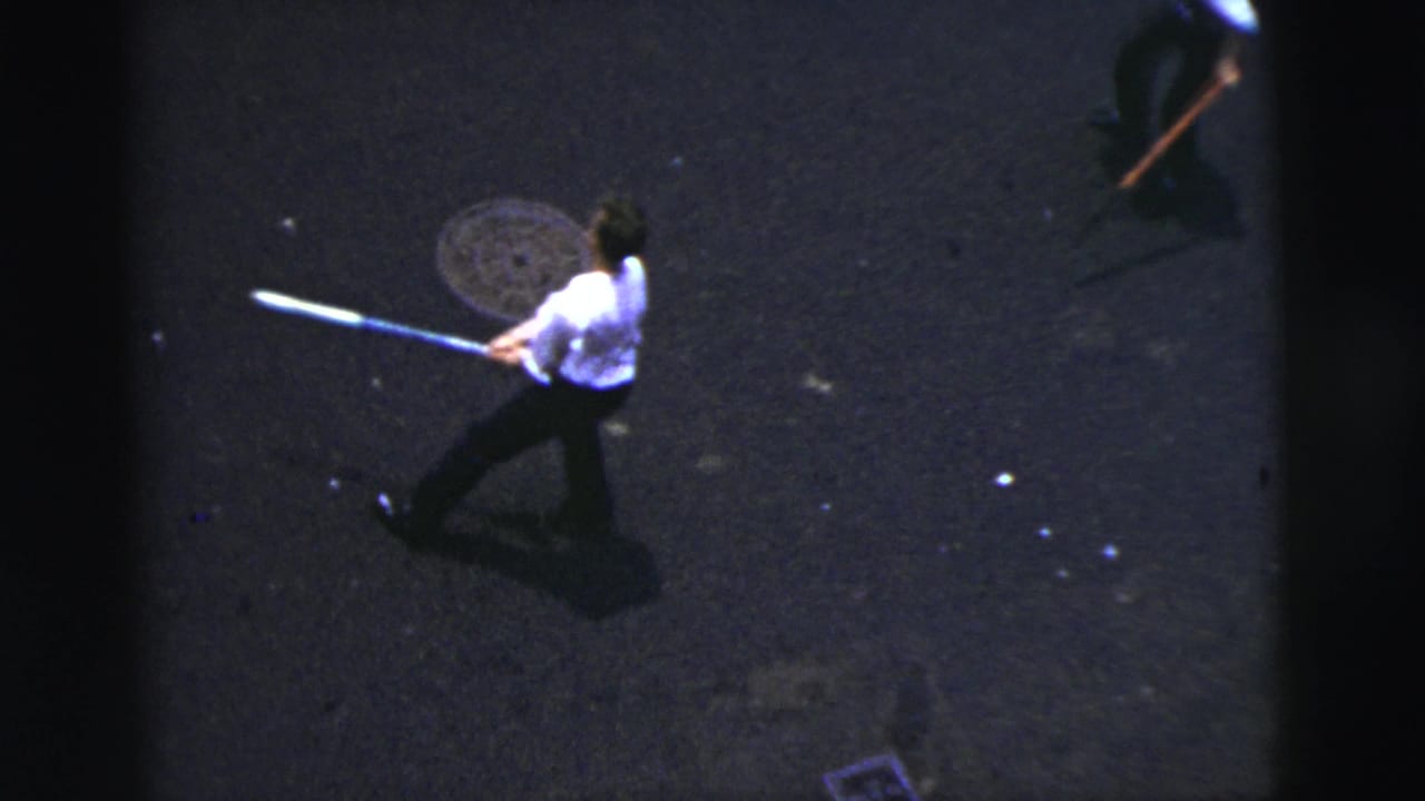 Man swings a bat across a dark street while another person is partly visible nearby.
