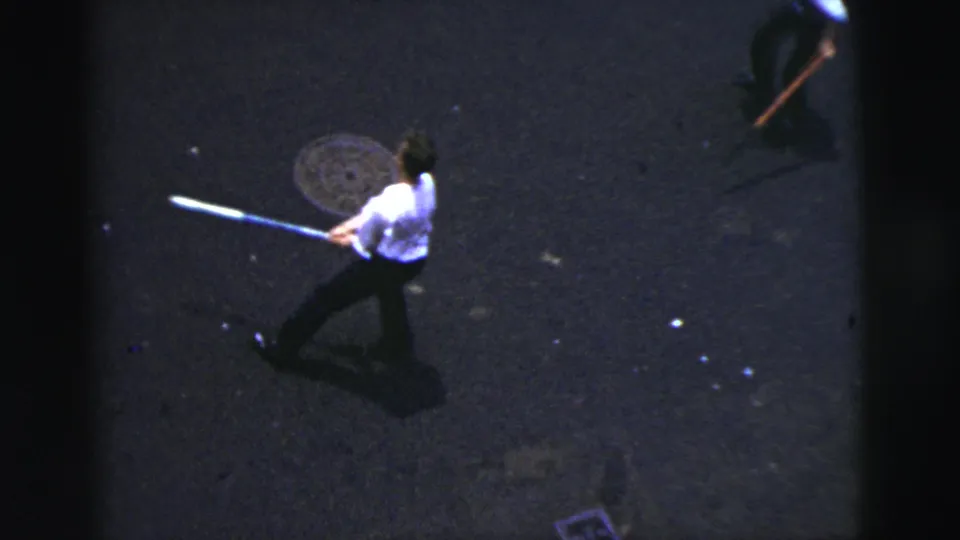 Elevated 1956 New York City home-movie frame showing a man swinging a bat on dark pavement with a partial passerby above.