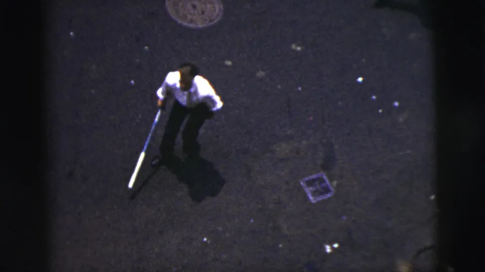 Elevated 1956 New York City home-movie view of a man pausing with a bat on dark asphalt beside a manhole cover and debris.