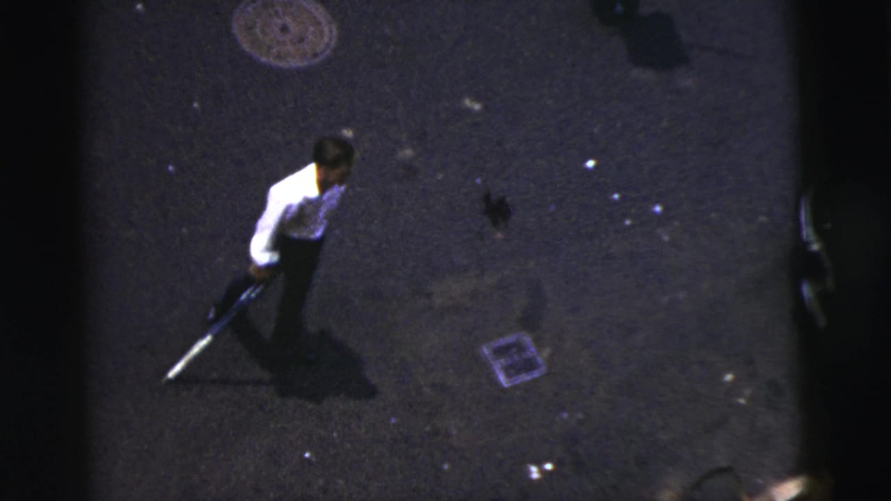 Man in a white shirt stands on dark asphalt holding a stick near a manhole cover.