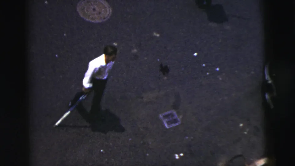 Elevated 1956 New York City home-movie view of a man in a white shirt holding a stick on a dark street.