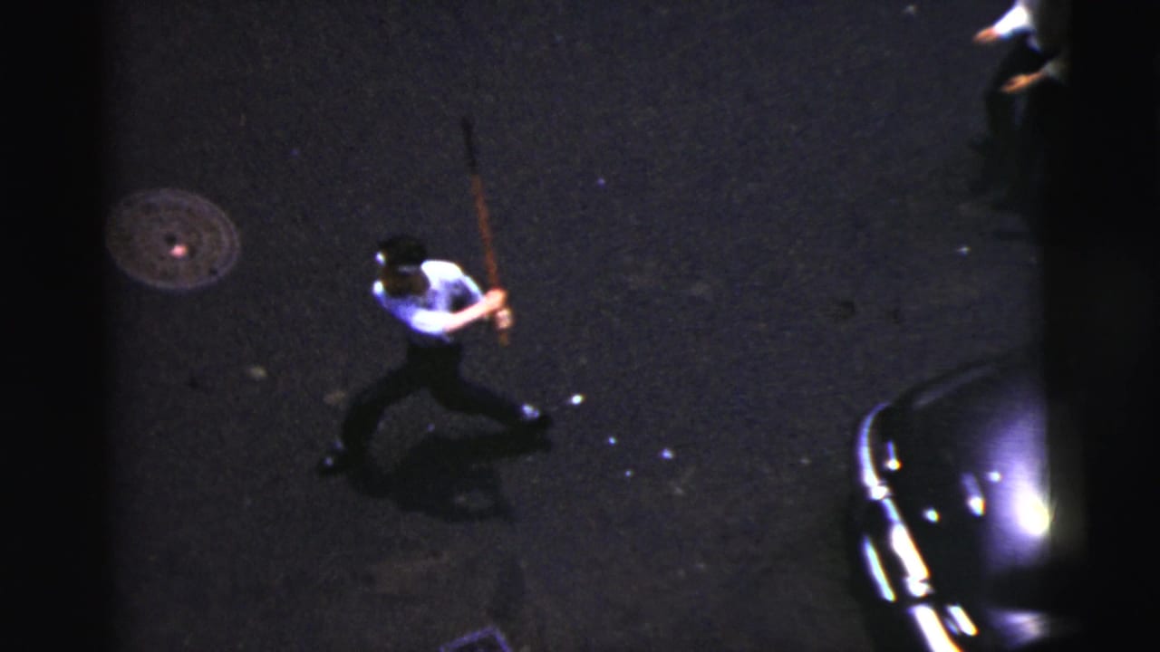Man runs or swings a stick on the street beside a parked car and manhole cover.