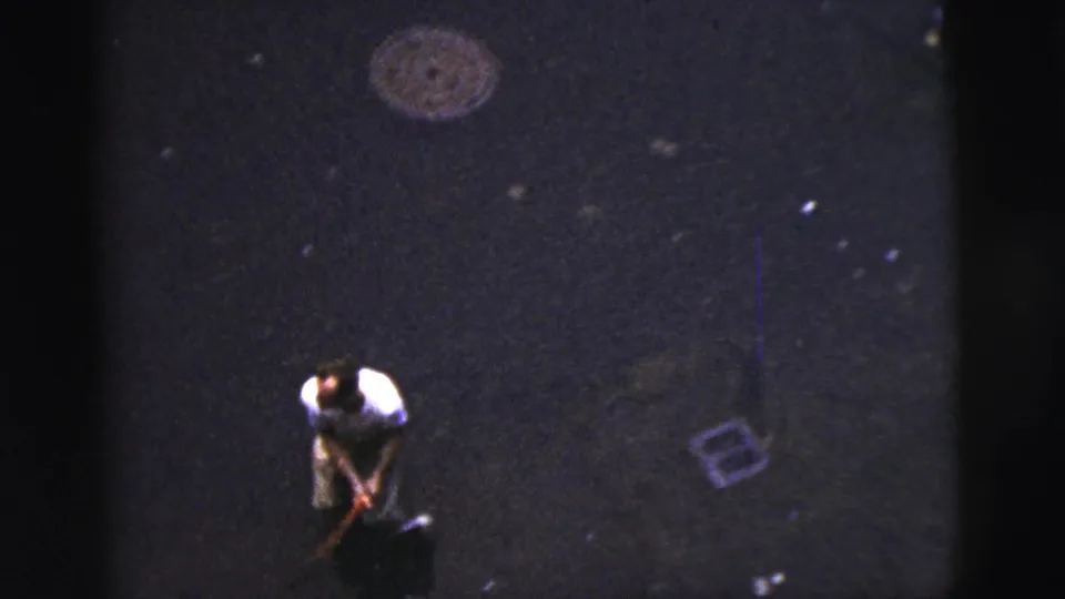 Elevated 1956 New York City home-movie view of an adult man on a dark paved street during an informal nighttime baseball game.