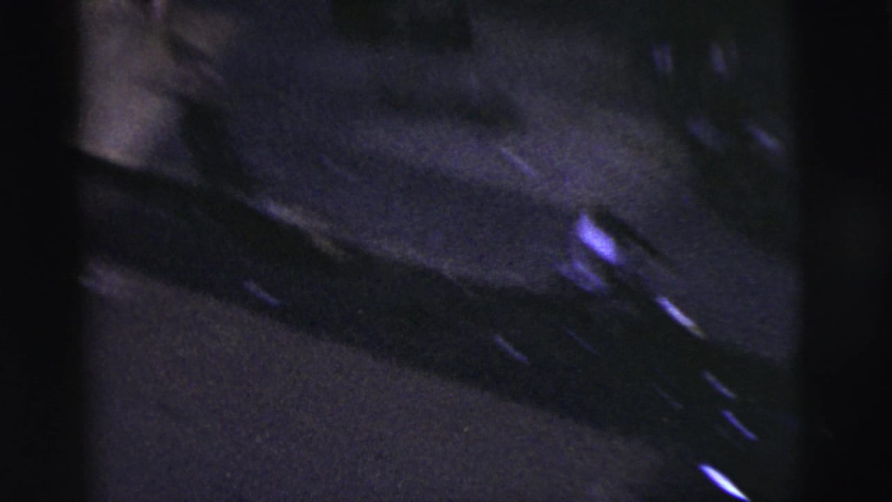 Dim 8mm frame from the New York City street game showing a parked car along the curb and dark pavement under low light.