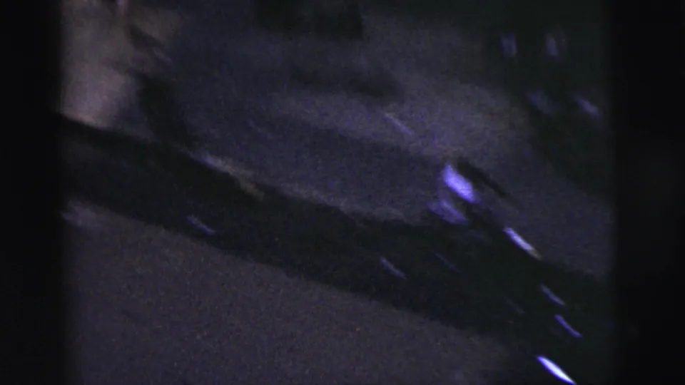 Dim 8mm frame from the New York City street game showing a parked car along the curb and dark pavement under low light.