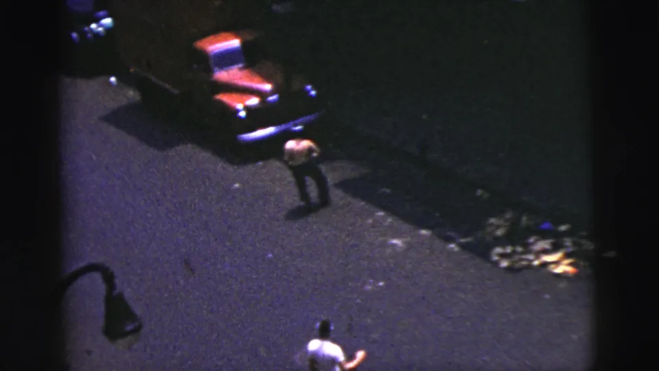1956 New York City home-movie frame showing an adult man in a white shirt with a bat during a nighttime street baseball game.