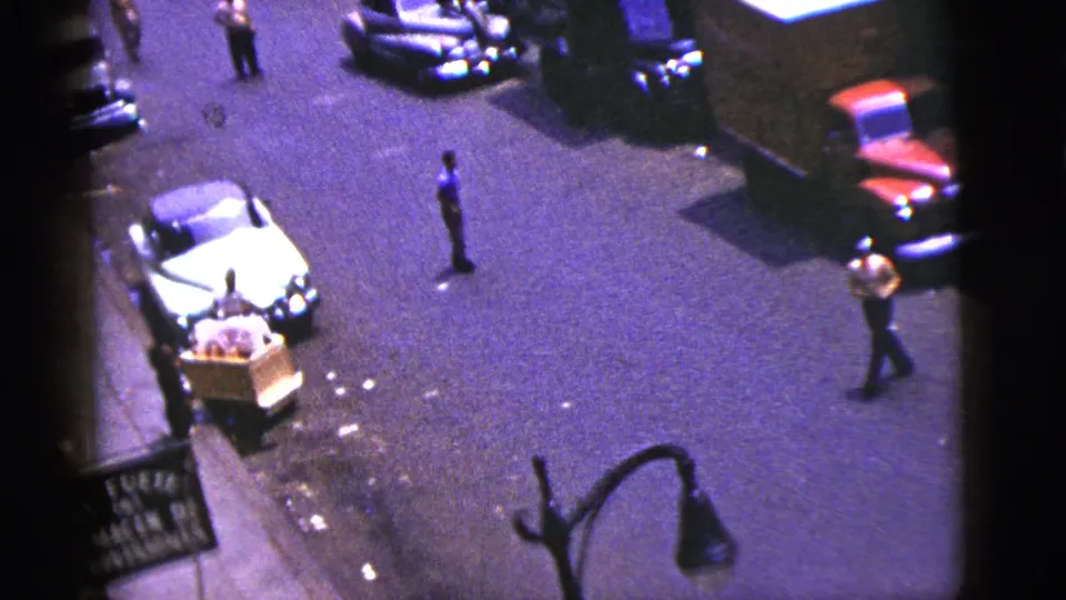 1956 New York City home-movie frame showing an elevated view of parked cars, pedestrians, a wooden pushcart, and a storefront sign.