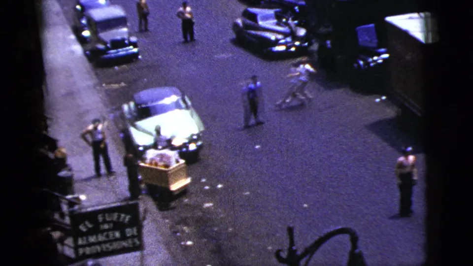 1956 New York City home-movie frame with pedestrians, classic parked cars, a curbside pushcart, and a Spanish-language grocery sign.