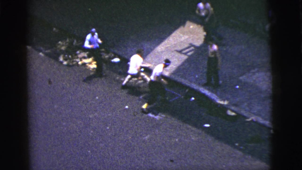 1956 New York City 8mm frame showing several men near a curb, scattered debris, and a tense sidewalk interaction from above.
