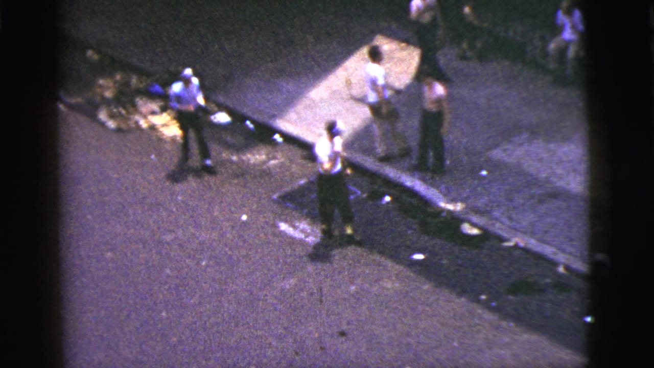 High-angle 1956 New York City street frame with men gathered near a curb and debris during a tense street moment.