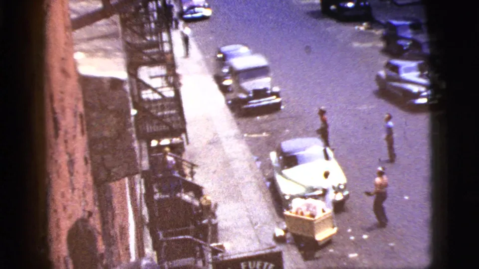 1956 New York City street frame with parked cars, building fire escapes, a pushcart, and people spaced along the roadway.