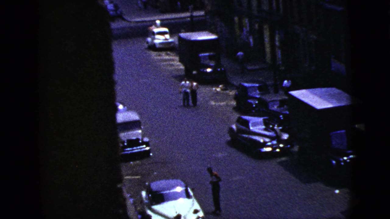 High-angle street view with parked cars, a truck, and several pedestrians in the roadway.