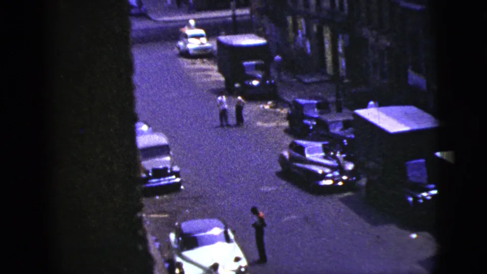 1956 New York City street scene from above, showing parked cars, a truck, and a person standing near the curb.