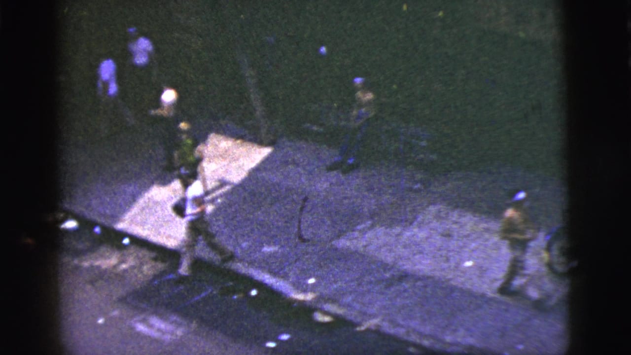High-angle home-movie frame shows pedestrians in 1950s New York City moving past a dim sidewalk corner.