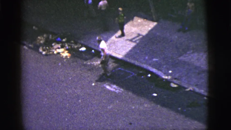 1956 New York City street frame with a pedestrian near visible curbside trash and a shadowed sidewalk.
