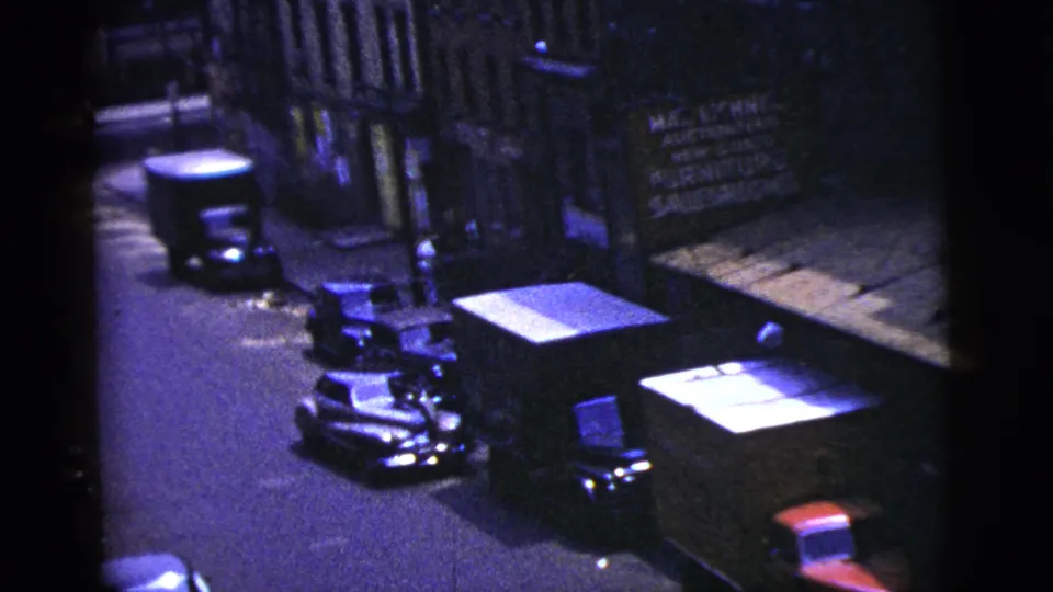 1956 New York City street view with parked cars, storefront facades, and a furniture salesroom sign visible from above.