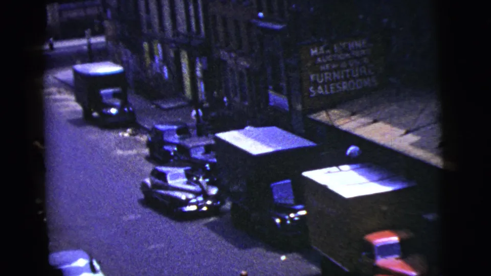 High-angle 1956 New York City home-movie frame showing parked cars, storefronts, and a small figure near the street edge.