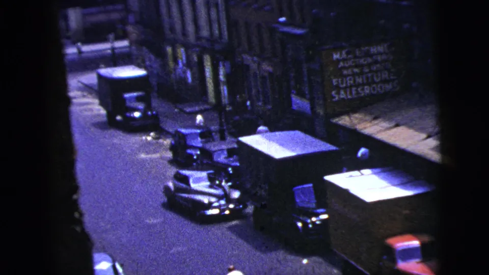 1956 New York City street frame with parked cars, storefronts, a sidewalk sign, and dark 8mm contact-sheet borders.