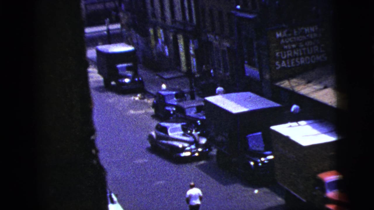 Elevated street view with parked cars, trucks, a storefront sign, and a man in a white shirt near the road.