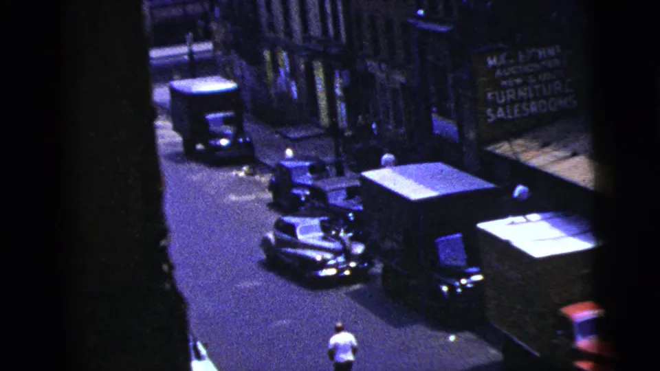 1956 New York City home-movie frame showing an elevated view of a street with parked vehicles, a furniture salesroom sign, and a man below.