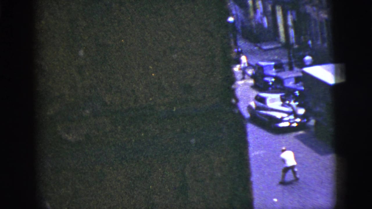 1956 New York City street frame with a man in a white shirt near parked cars, partly obscured by dark building edges and film exposure.