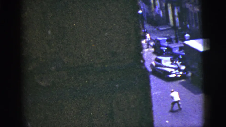 1956 New York City street frame with a man in a white shirt near parked cars, partly obscured by dark building edges and film exposure.