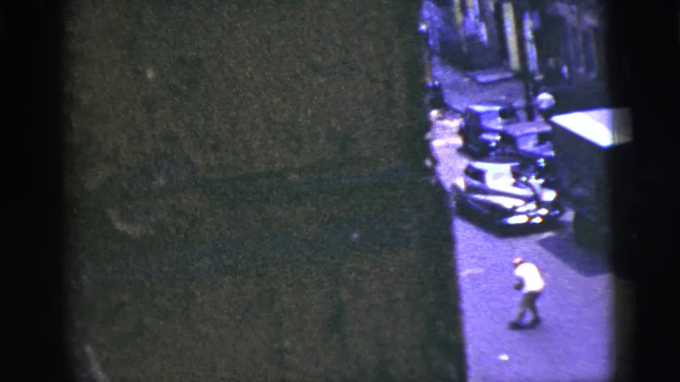 1956 New York City home-movie frame showing a man reaching in the roadway near parked cars, with heavy shadow and soft 8mm detail.