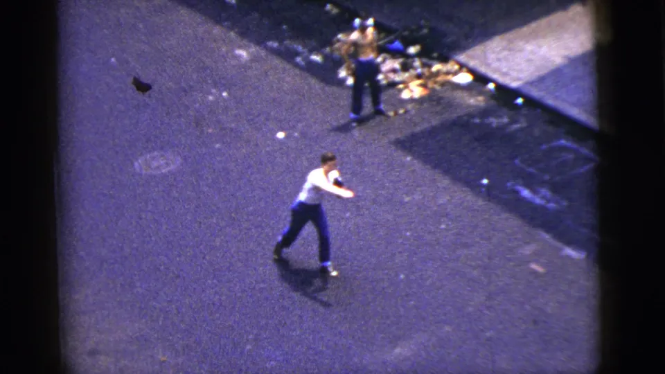 1956 New York City frame showing two men in the street during a catch game, with sidewalk debris, shadow, and open roadway visible.
