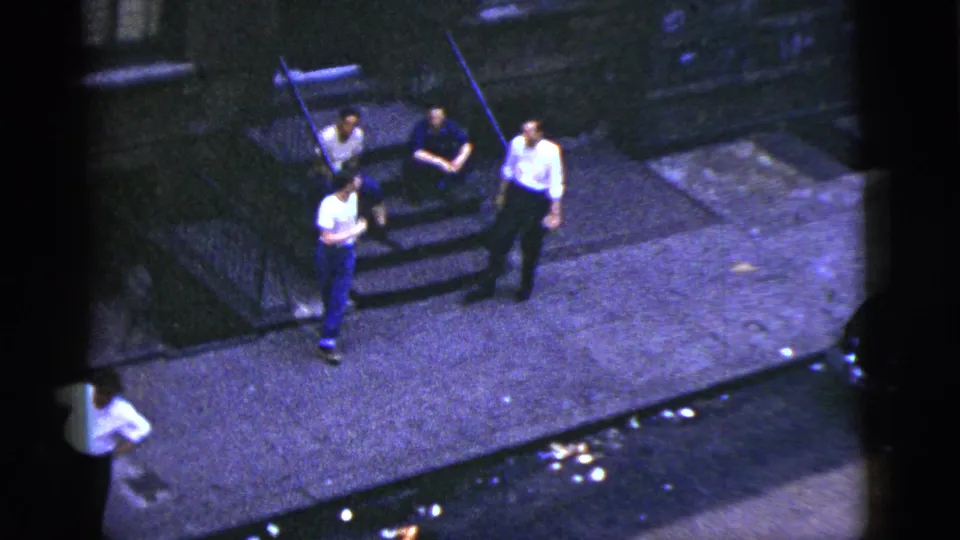 Four young men stand and sit near building steps on a New York City sidewalk in 1956 home-movie footage.