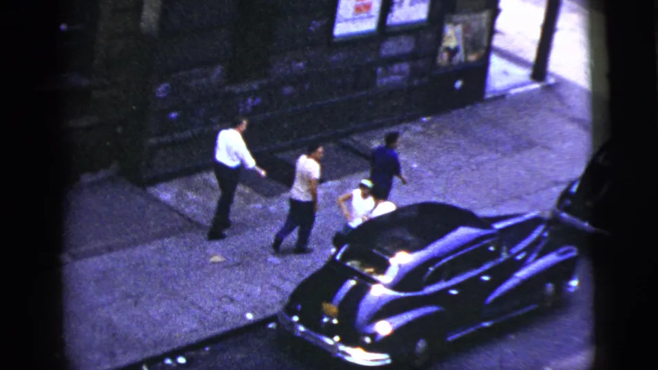 Young men walk beside a dark sedan on a New York City sidewalk, with building posters and a doorway in the background.