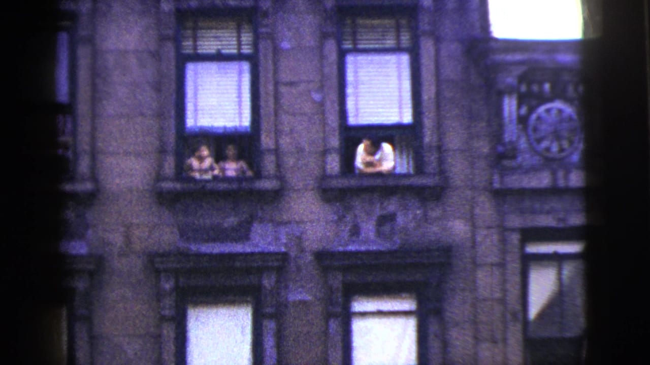 1956 New York City home-movie frame showing a stone apartment facade with people leaning or sitting at upper windows.