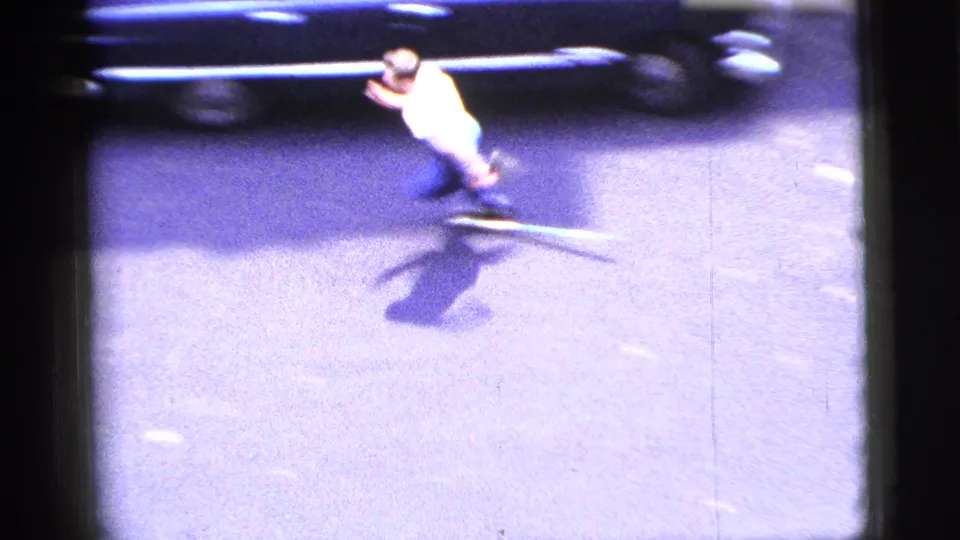 1956 New York City home-movie frame showing a man in a white shirt and blue pants running across a street beside a dark car.