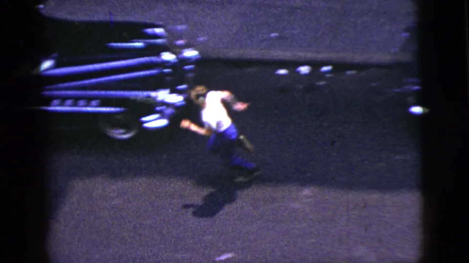 1956 New York City frame of a man in a white shirt running near a dark mid-century car on a shadowed street.