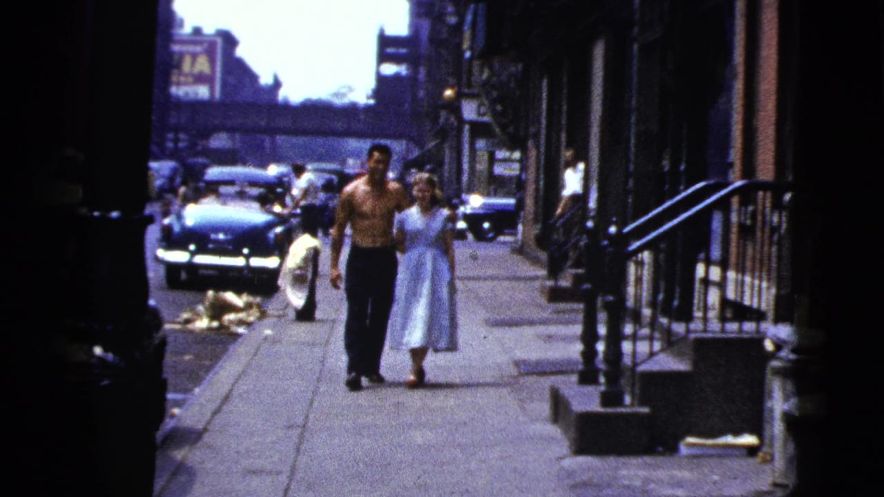 In 1956 New York City footage, a shirtless man and a woman in a light blue dress walk toward the camera on a sidewalk.