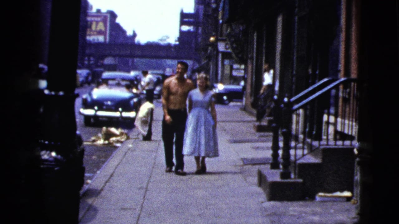 The man and woman walk arm in arm along a New York City sidewalk, with cars, pedestrians, and an elevated structure behind them.