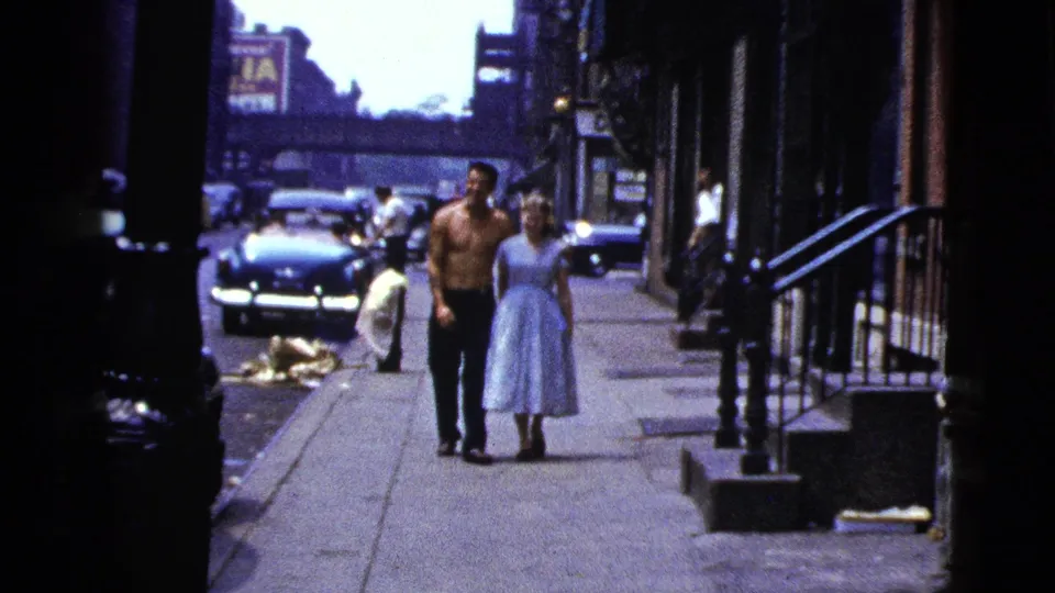 The man and woman walk arm in arm along a New York City sidewalk, with cars, pedestrians, and an elevated structure behind them.