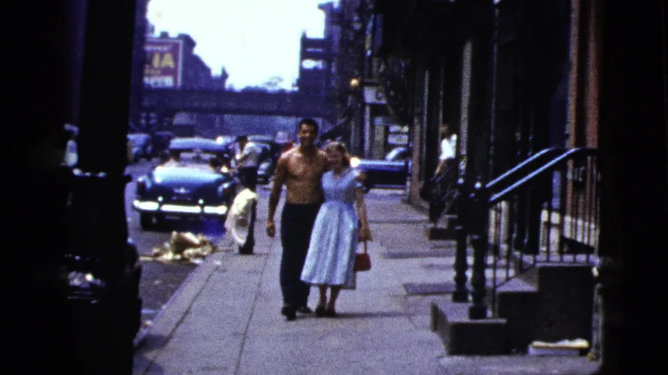 The couple is closer in frame, walking arm in arm past stoops and storefronts on a 1950s New York City sidewalk.