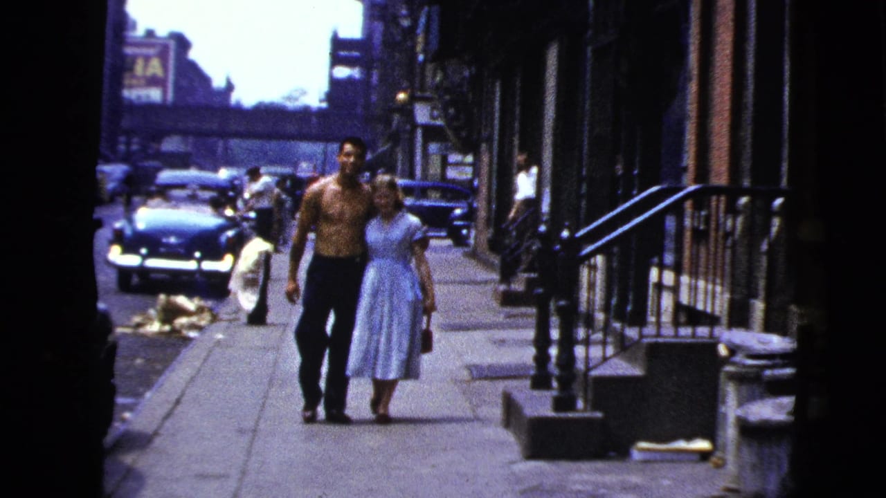 A shirtless man and a woman in a light dress walk arm-in-arm down a city sidewalk.
