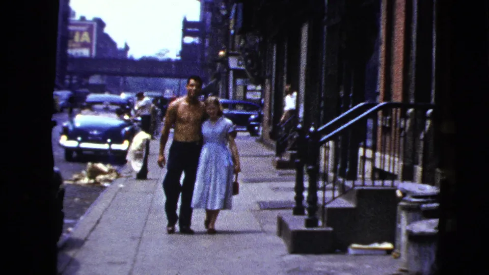 In New York City, 1956, a shirtless man and a woman in a light blue dress walk toward the camera past stoops and older cars.