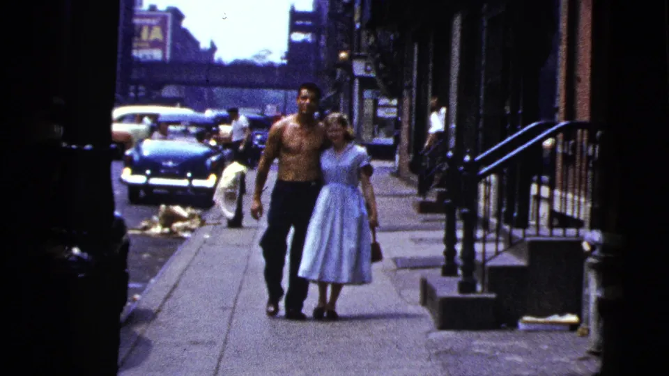 The couple approaches in soft 8mm color, with a dark car, sidewalk litter, stoops, and building fronts visible behind them.